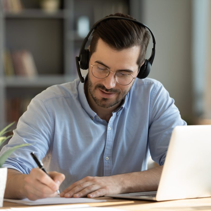 Focused 35s man sit at desk wear headphones watch webinar use laptop gain new knowledge writing notes. Video conference communication, negotiation remotely, study online, e-learn, self-educate concept